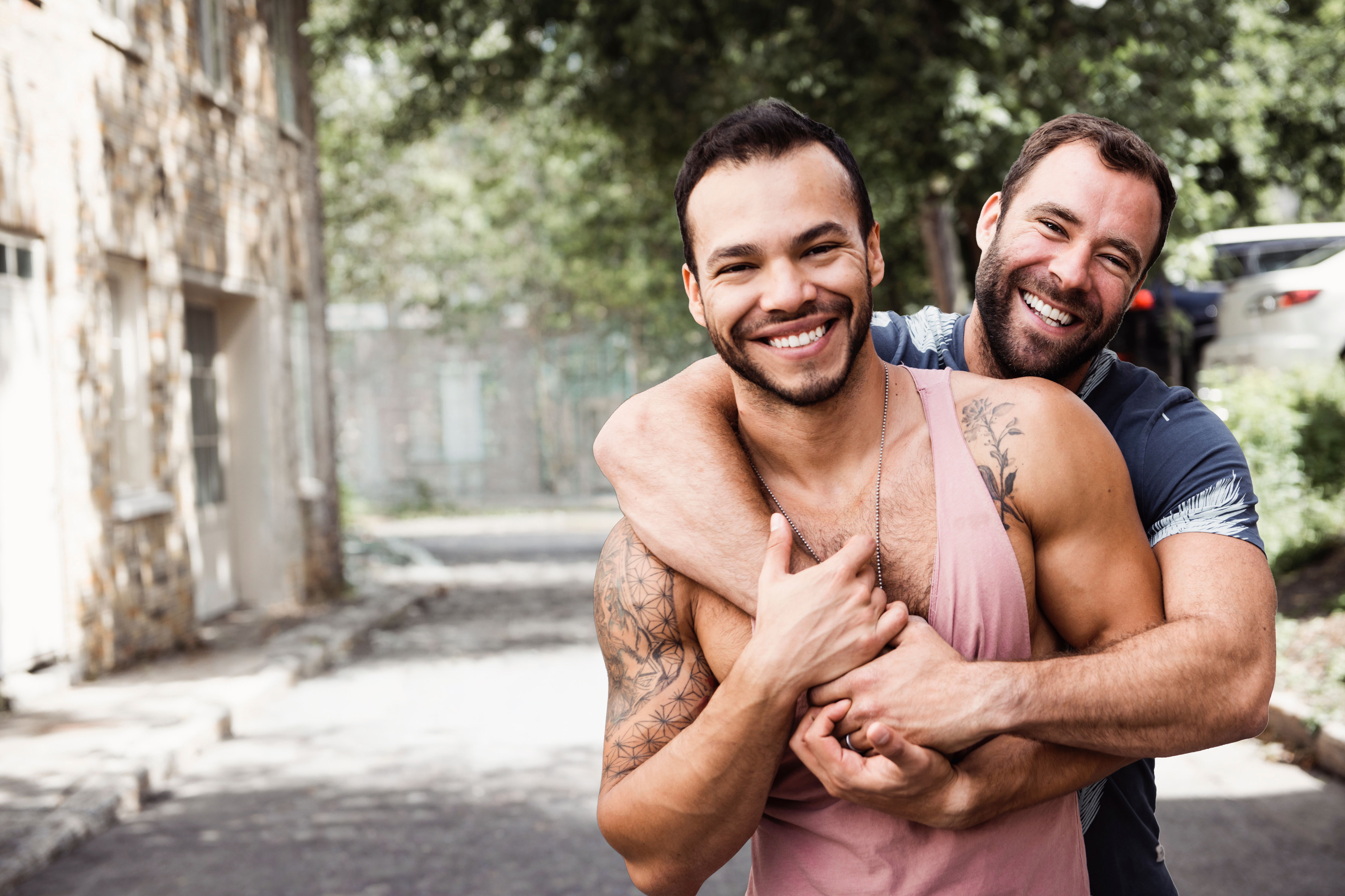 Two smiling men embrace outdoors on a tree-lined street with a blurred city background.