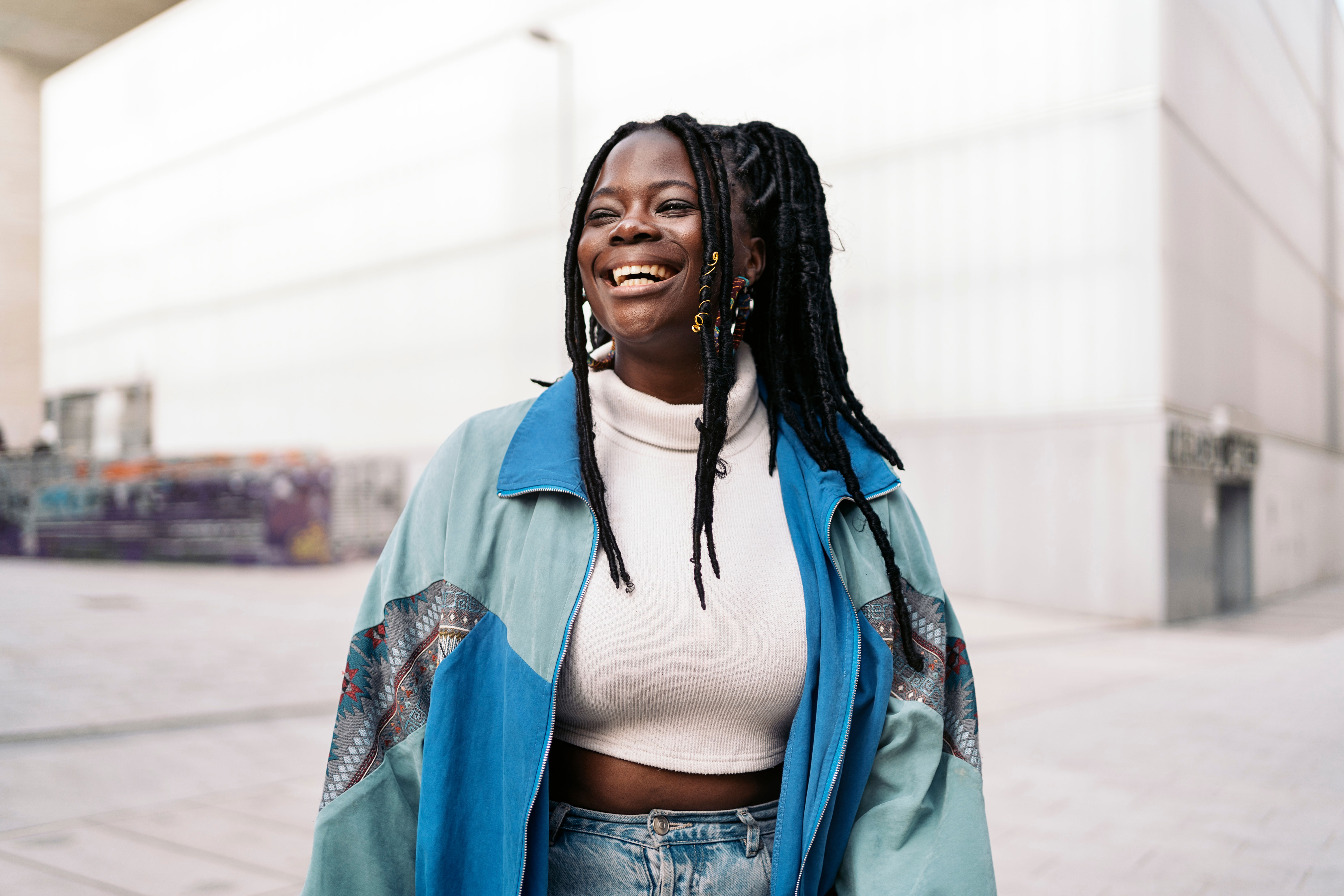 An exuberantly smiling Black woman with long, braided hair, wearing a blue jacket and a white turtleneck top, outdoors against a light urban background.