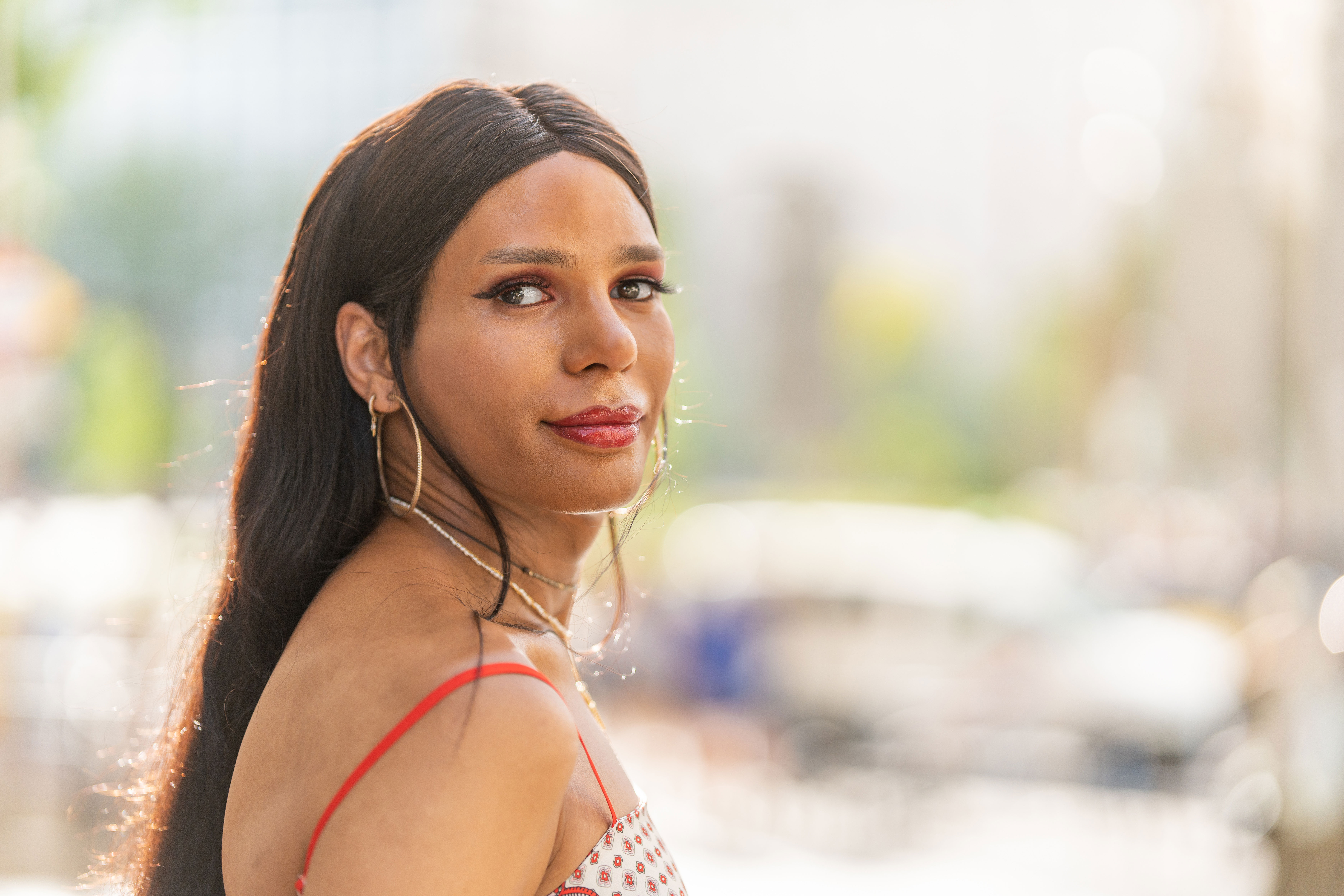 Portrait of a woman with long dark hair wearing hoop earrings and a patterned red-strap top, looking over her shoulder outdoors during the day.