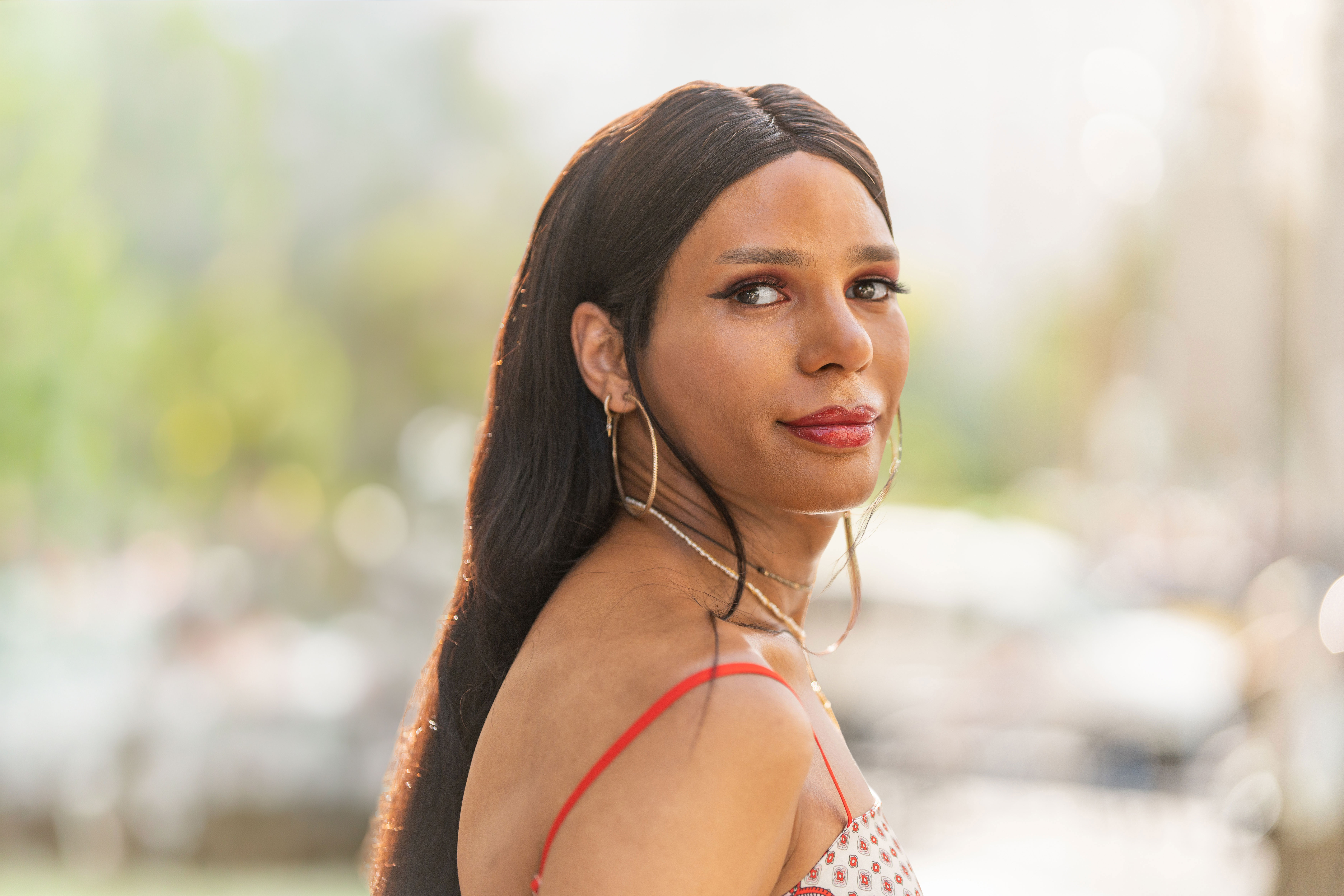 Portrait of a woman with long dark hair wearing hoop earrings and a patterned red-strap top, looking over her shoulder outdoors during the day.